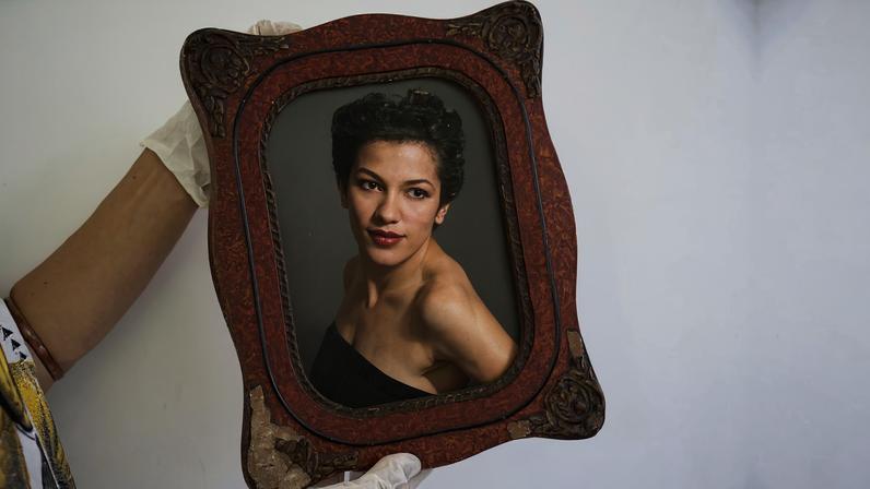 Braços com mãos enluvadas exibem porta-retratos com foto preta-e-branca de uma jovem mulher negra maquiada, usando um vestido tomara-que-caia