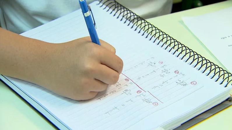 Mão de uma pessoa escrevendo com uma caneta em um caderno de espiral, preenchido com exercícios matemáticos dispostos em colunas e anotações numeradas
