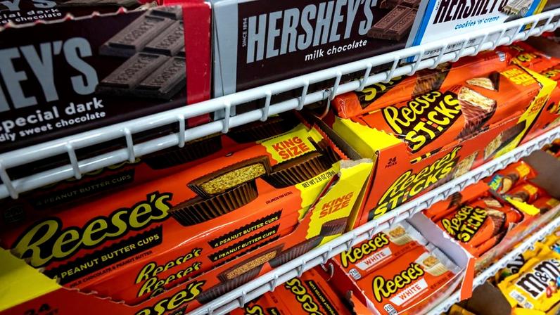 Doces variados, incluindo barras Hershey's e Reese's, expostos em prateleiras de metal de uma loja.