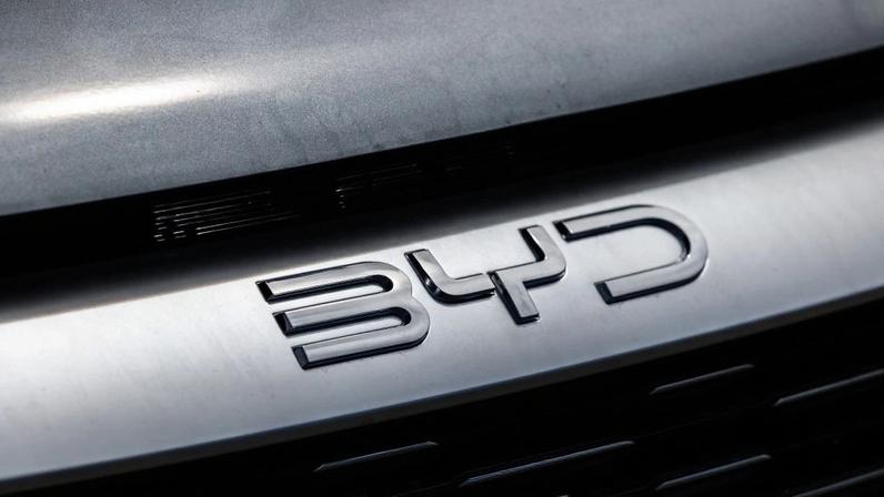 Logo metálico e brilhante da BYD na grade frontal de um carro cinza.