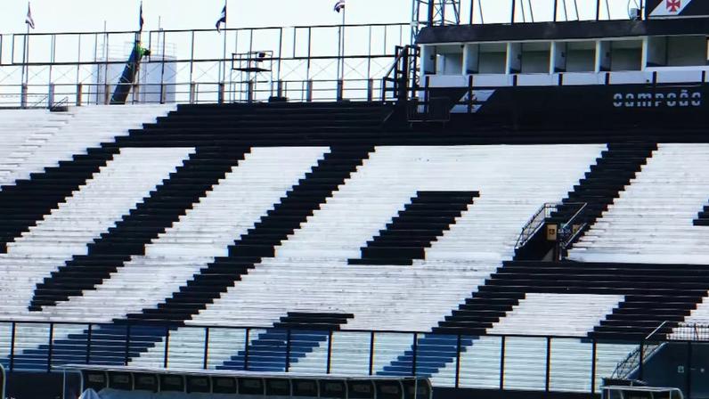 Arquibancada de estádio com cadeiras pretas e brancas formando a palavra “VASCO”. Acima, estrutura com bandeiras e inscrição “CAMPEÃO”.