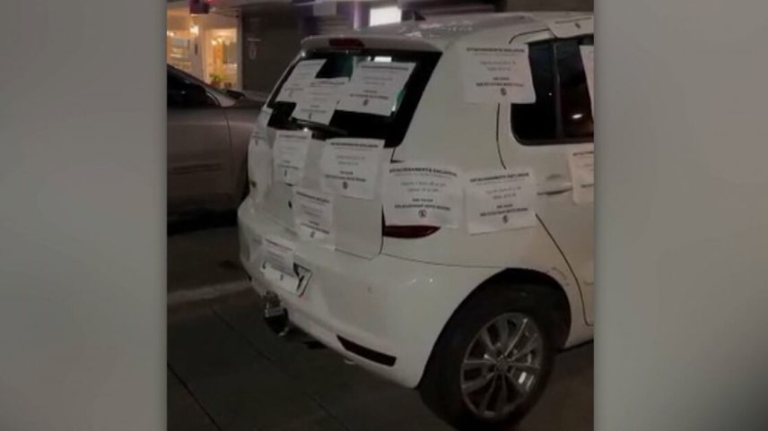 A imagem mostra a traseira de um carro branco estacionado à noite em uma área urbana iluminada artificialmente. Toda a parte traseira e lateral do veículo está coberta por diversas folhas de papel impressas, coladas sobre a lataria e o vidro. Ao redor do veículo, outros carros estão estacionados, e vitrines iluminadas de estabelecimentos podem ser vistas ao fundo, sugerindo que o veículo está parado em um estacionamento externo próximo a lojas ou serviços.