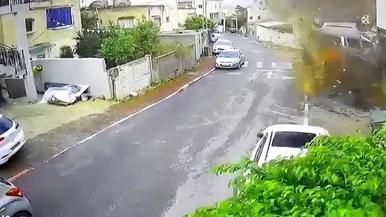 Câmera de segurança registra uma rua residencial estreita, com carros estacionados ao longo da via. No canto direito da imagem, um veículo aparece no ar, virado de cabeça para baixo, no momento de uma explosão que levanta poeira e destroços