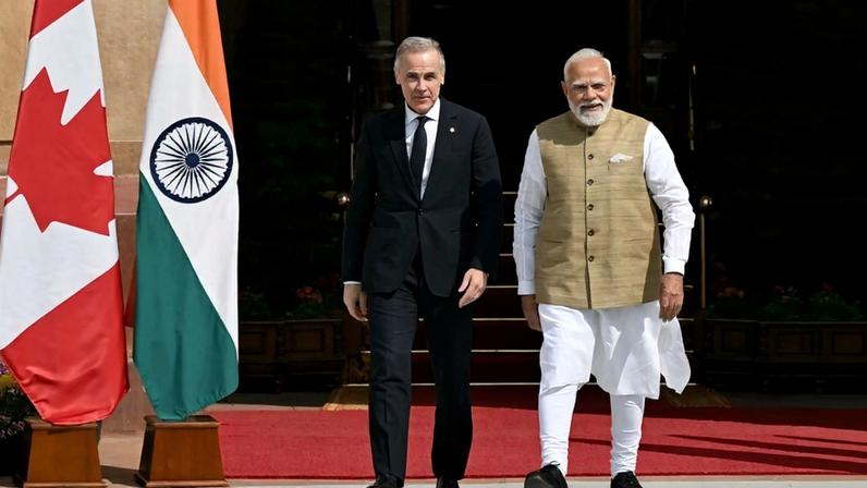 Carney and Modi walk side-by-side near Indian and Canadian flags. Both men smile slightly.