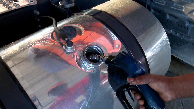 Pessoa segura bico de combustível azul encaixado em tanque metálico cilíndrico e brilhante de caminhão. Cena retrata ato de abastecimento de veículo grande em ambiente de transporte e logística.