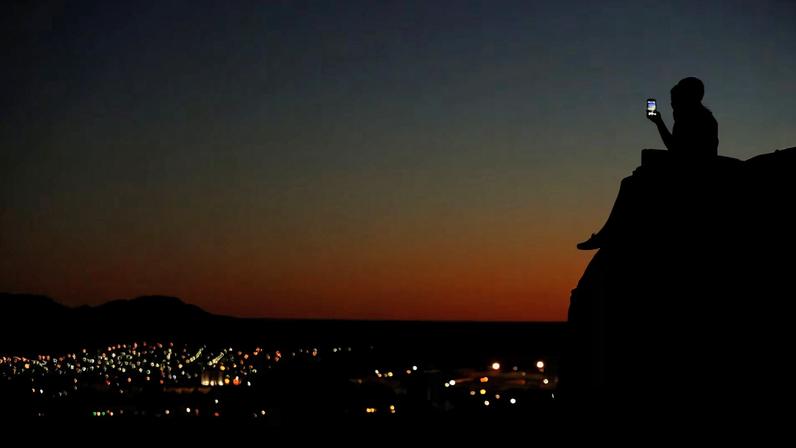 A young couple look at their phone as they sit on a hillside after sun set in El Paso, Texas