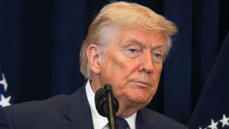 Donald Trump usando terno escuro e gravata, posicionado atrás de um microfone, com duas bandeiras estreladas ao fundo sobre um painel azul‑escuro