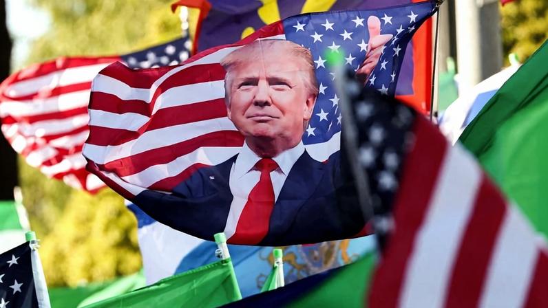 An array of flags are seen billowing in the wind, including Iranian flags, American flags, and a large American flag embellished with a cut-out photo of Donald Trump giving a thumbs up.