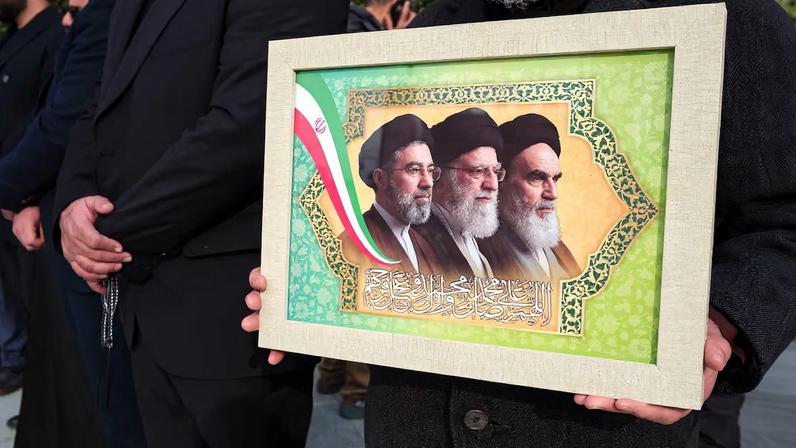 Un asistente al entierro del almirante Ali Shamjani este sábado en Teherán porta una imagen del nuevo líder supremo de Irán, Mojtaba Jameneí; su antecesor y padre, Alí Jameneí, y el fundador de la República Islámica, el ayatolá Jomeiní.