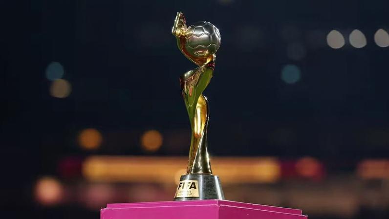 Troféu dourado da Copa do Mundo Feminina da Fifa está sobre pedestal rosa. A taça tem figura estilizada segurando bola no alto e base com placa gravada “FIFA WOMEN'S WORLD CUP”.