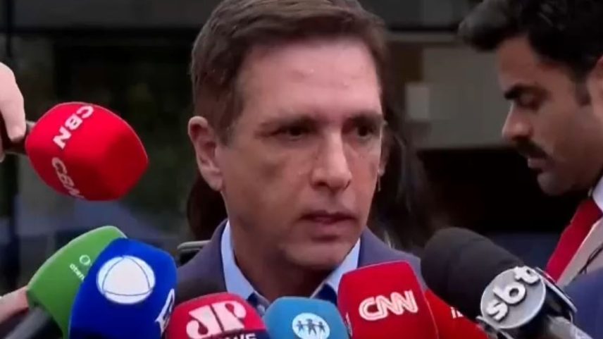 Homem falando diante de vários microfones de diferentes emissoras de TV e rádio, em uma coletiva de imprensa. Ao lado, outro homem com fone no ouvido acompanha a cena.