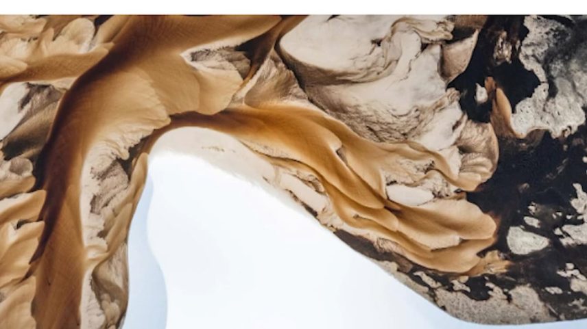 Vista aérea de um deserto com dunas de areia claras e formações rochosas escuras. As curvas suaves das dunas contrastam com as texturas ásperas das pedras, que apresentam manchas brancas, lembrando depósitos minerais.