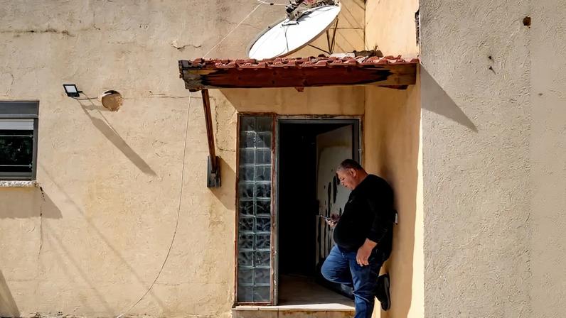 A man in a black shirt and blue jeans checks his cellphone outside of a tan house in northern Israel.