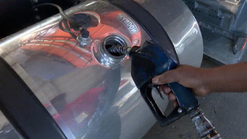 Tanque metálico de combustível sendo abastecido. Uma mão segura uma bomba de combustível azul inserida na abertura do tanque cilíndrico prateado
