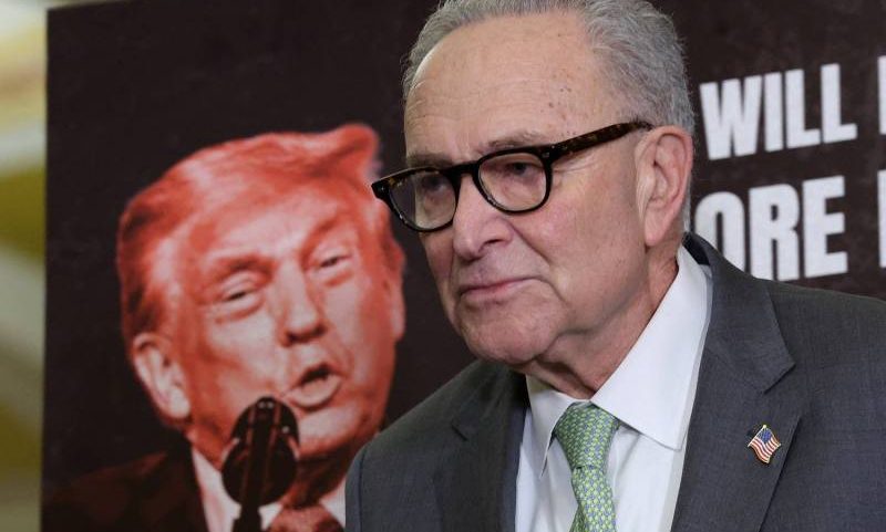 U.S. Democratic Sen. Charles Schumer speaks into a microphone while standing in front of a large poster of U.S. President Donald Trump.