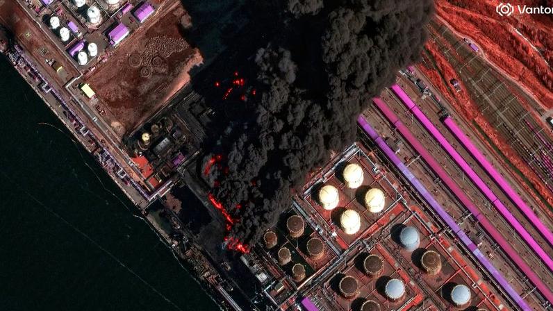 Incêndio em refinaria de petróleo com fumaça preta e estruturas industriais coloridas ao redor