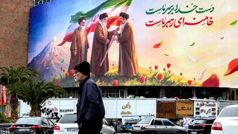 A man in a black jacket and beanie walks underneath a banner with an illustration displaying a transfer of the Iranian flag between the three supreme Leaders.
