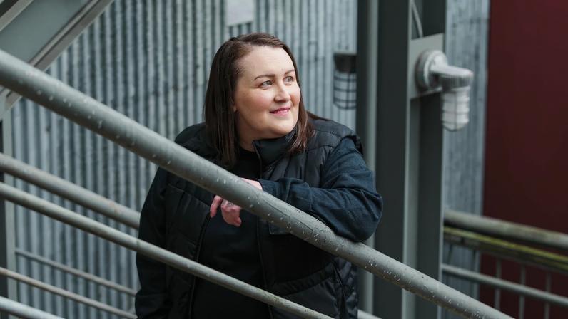 Kyla Gillespie in a black jacket standing on a metal staircase in an industrial setting, looking to the side.