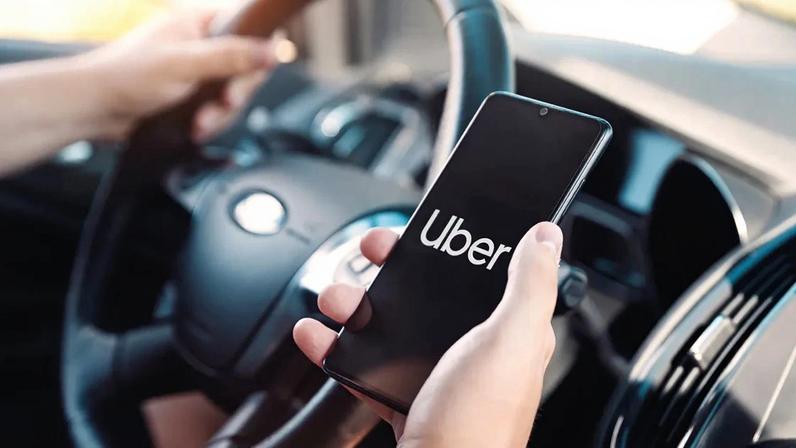 IMagem de um motorist segurando o volante com a mão esquerda e com um celular mostrando o logo da Uber na direita