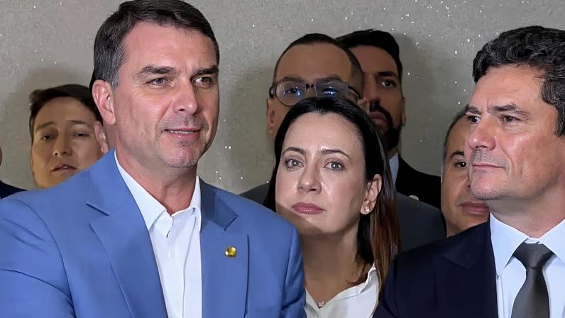 Grupo de pessoas em ambiente interno, todas em trajes formais. Em primeiro plano, dois homens de terno, um com paletó azul claro e camisa branca, outro de terno escuro com gravata.