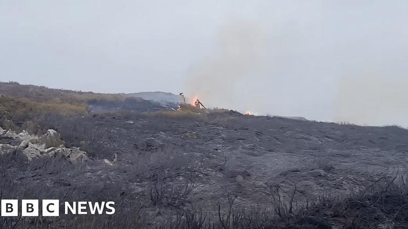 An area of gorse and brash on a slope, with flames burning in the distance. A figure can be seen silhouetted against the flames. It is daylight, on a cloudy day