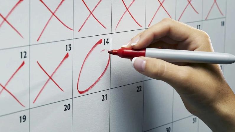 Imagem de uma mão segurando uma caneta vermelha enquanto circula a data 14 em um calendário de papel com fundo branco e números pretos. Datas 12 e 13 têm xis vermelhos. Imposto de Renda