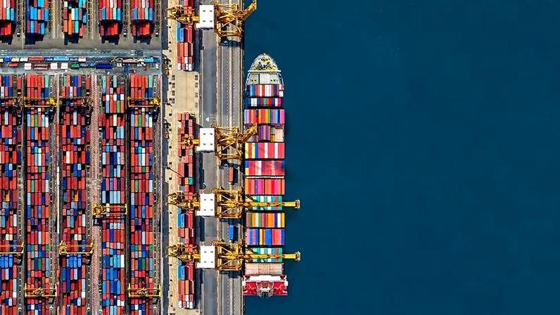 Vista aérea de um porto com contêineres coloridos organizados à esquerda, guindastes amarelos no centro-esquerdo, um navio cargueiro atracado à direita e o mar azul profundo ao fundo.