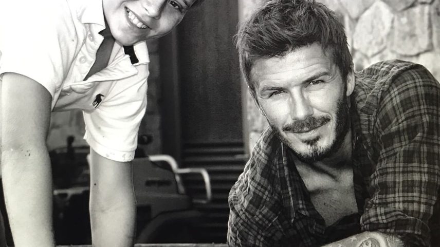 David Beckham and a young Brooklyn Beckham smiling and leaning over a barrel.