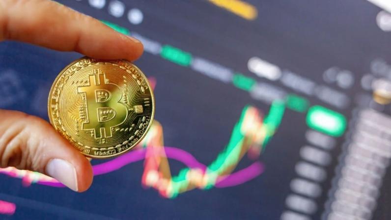 dedos seguram moeda de Bitcoin à frente de gráfico financeiro