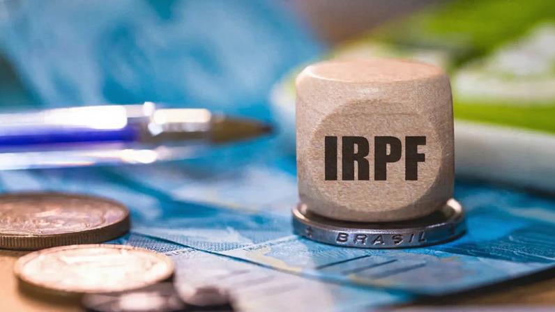 um peso de papel com as siglas IRPF em cima de uma moeda e de cédulas de real para ilustrar reportagem sobre ir 2026 e declaração do imposto de renda 2026