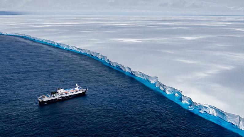 A ship sails towards a towering ice shelf with a bright blue band at its base where it meets the sea, under a cloudy polar sky.