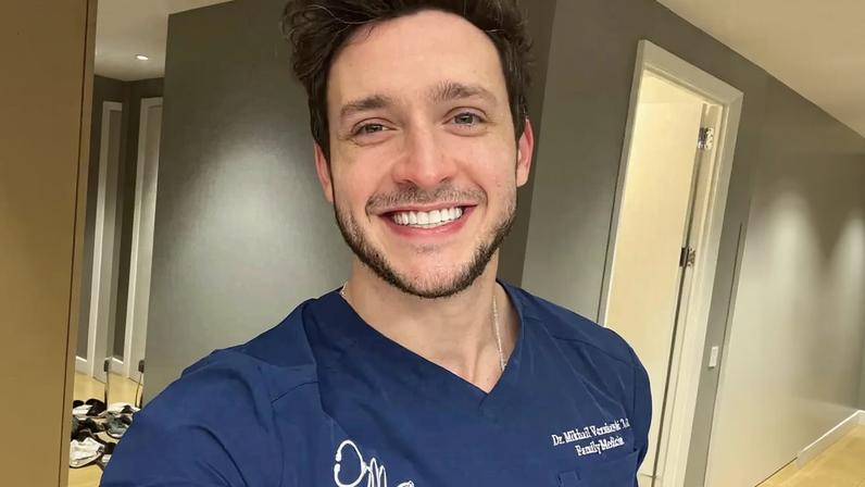 Portrait of Dr. Mikhail Varshavski, a family medicine doctor, smiling in blue scrubs.