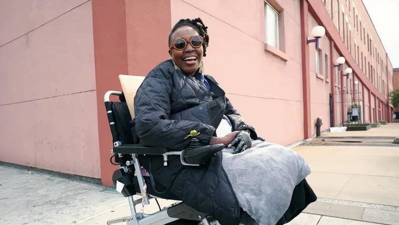 Seated in an electric wheelchair, a person wears a black quilted coat and brown-framed sunglasses. They are smiling broadly with their mouth open, against a pink building.