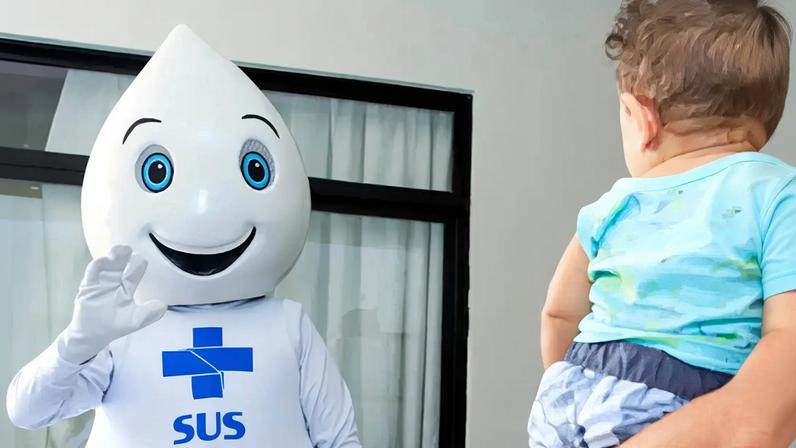 Foto do Zé Gotinha acenando para um bebê no colo da mãe.