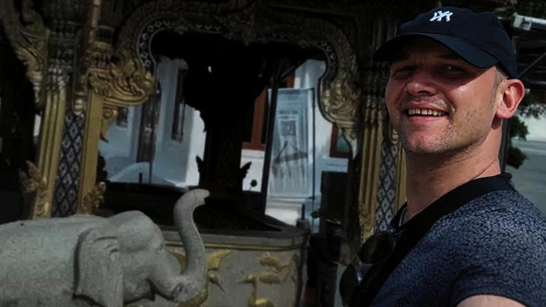 Rory McColl , who is wearing a black baseball cap, takes a selfie in front of a stone elephant and a historic building