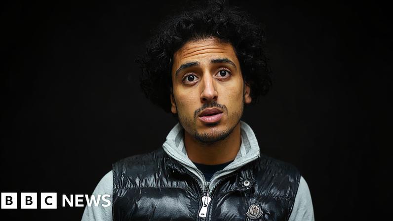 A young man called Akwa, captured by secret filming, stands inside a shop, in front of shelves with rows of colourful packaging for vapes. He wears a shiny black bodywarmer with a grey top underneath, he has curly short dark hair and he looks to be in conversation.