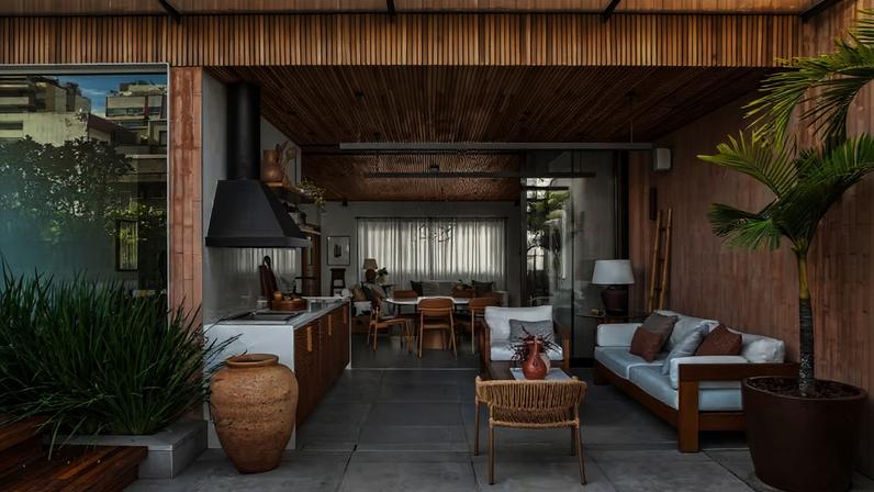 AMBIENTES INTEGRADOS | Na cobertura, varanda com churrasqueira, piscina e living são totalmente conectados, tendo como pano de fundo materiais naturais e atemporais. Essa continuidade visual é reforçada pelo teto de madeira ripada, executado pela Targa Engenharia, e pelo piso de porcelanato Burlington Stone, inspirado na pedra natural de mesmo nome, originária da Inglaterra, adquirido na Ekko Revestimentos, loja que também forneceu os tijolinhos na parede da varanda, onde estão sofá, poltrona e mesa de centro da Franccino Móveis. Almofadas da Galeria Hathi. O grande vaso marrom no piso é de Beatriz Santiago Paisagismo, com palmeira