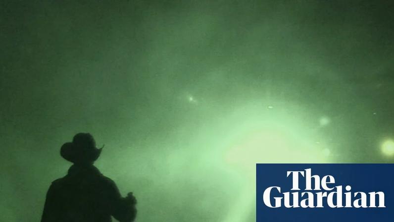 A night-vision camera shows a silhouette of a man holding a torch and looking at a bright light in the sky