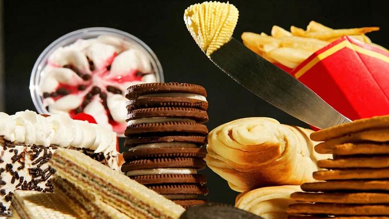 Diversos alimentos ultraprocessados dispostos juntos, incluindo biscoitos recheados empilhados, batatas fritas em embalagem vermelha, bolos com cobertura de chantili e confeitos, wafer com camadas de creme, pão enrolado e biscoitos redondos. Uma faca com manteiga é visível sobre o pão enrolado.