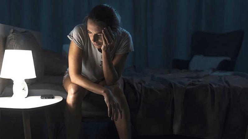 Mulher sentada na beira da cama em quarto escuro, iluminada por abajur branco sobre mesa lateral. Ela apoia a cabeça na mão, olhando para baixo, com controle remoto ao lado no móvel.