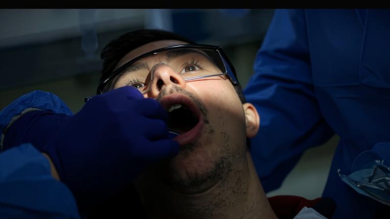 Paciente de óculos de proteção com boca aberta recebe exame odontológico. Profissional usa luvas roxas e uniforme azul, com foco de luz na boca do paciente.
