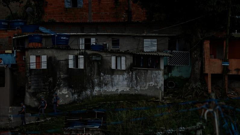 Residências simples de alvenaria com paredes desgastadas e telhados variados em área urbana. Vegetação rasteira e cercas de arame aparecem em primeiro plano, com árvores ao fundo. É possível ver três policiais com uniformes cinza descendo pela grama à esquerda da imagem