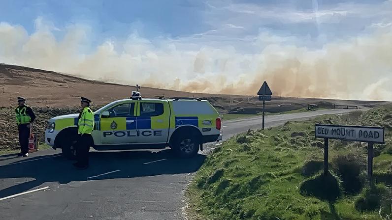 Side view of a West Yorkshire Police vehicle parked across a moorland road with two officers standing beside it. In the background a plume of brown and white smoke drifts across the moorland, partially obscuring an otherwise blue sky.