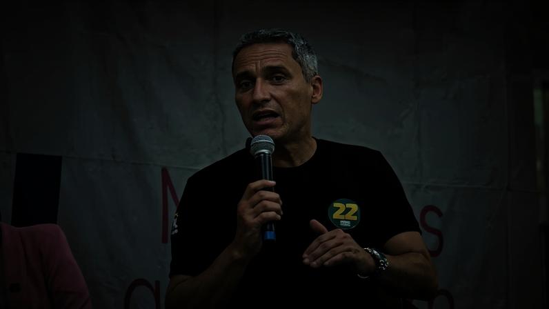 Gayer, homem com cabelos levemente grisalhos e vestindo uma camiseta preta, discursa com o microfone na mão