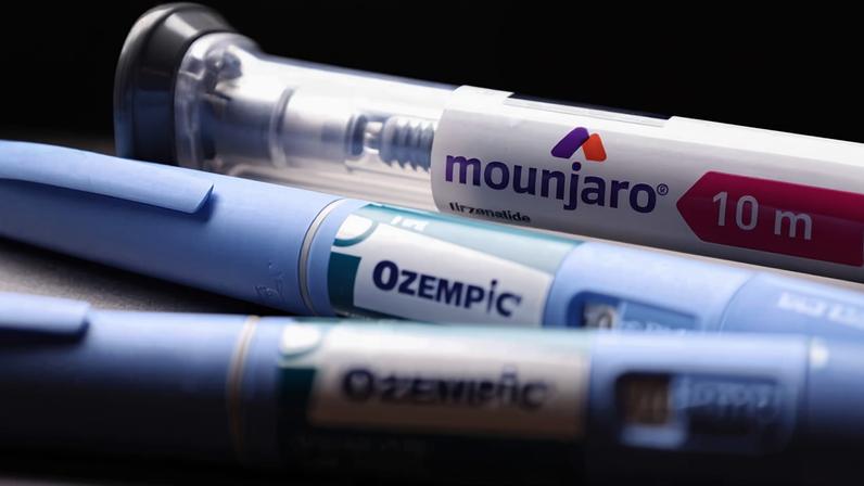 A imagem mostra três canetas de medicamento em primeiro plano. A caneta superior é transparente e contém o nome "mounjaro" e a dosagem de "10 mg". As duas canetas abaixo são azuis e têm o nome "Ozempic" visível. O fundo da imagem é escuro, destacando as canetas.