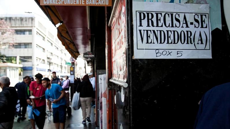 A imagem mostra um anúncio fixado em uma parede que diz "Precisa-se vendedor(a)" e "Box 5". Ao fundo, há pessoas caminhando na calçada, algumas carregando sacolas. O ambiente é de uma área comercial.