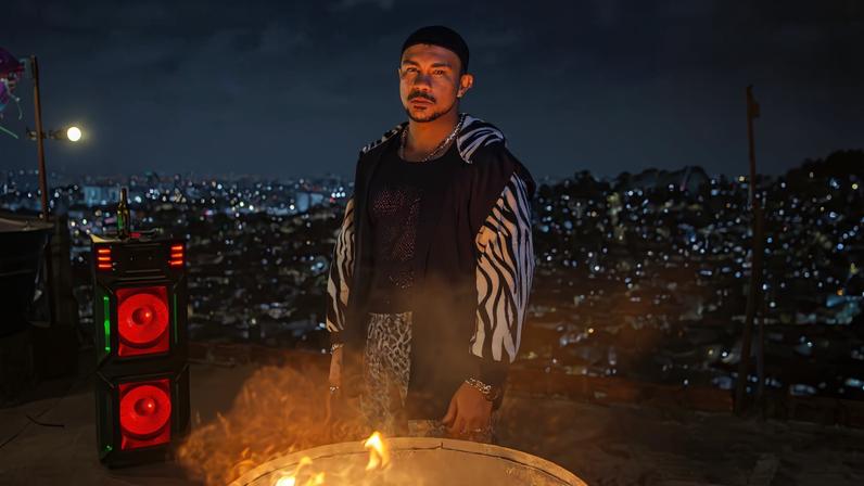 Homem jovem está em área externa à noite, próximo a uma fogueira em um tambor metálico. Ele veste moletom com capuz de estampa zebrada e olha para a câmera. Ao fundo, há uma cidade iluminada com muitas luzes e um equipamento de som com alto-falantes vermelhos acesos.