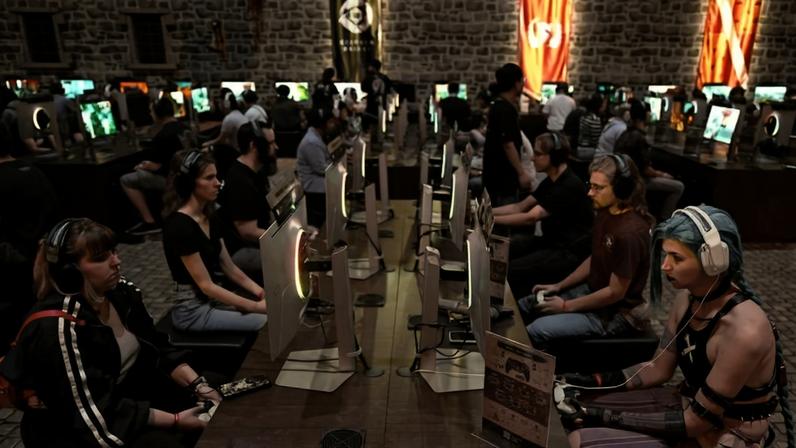 Dezenas de jogadores sentados em duas fileiras opostas em frente a monitores, competindo em videogames. O ambiente tem iluminação azul e paredes de pedra, com bandeiras penduradas ao fundo.