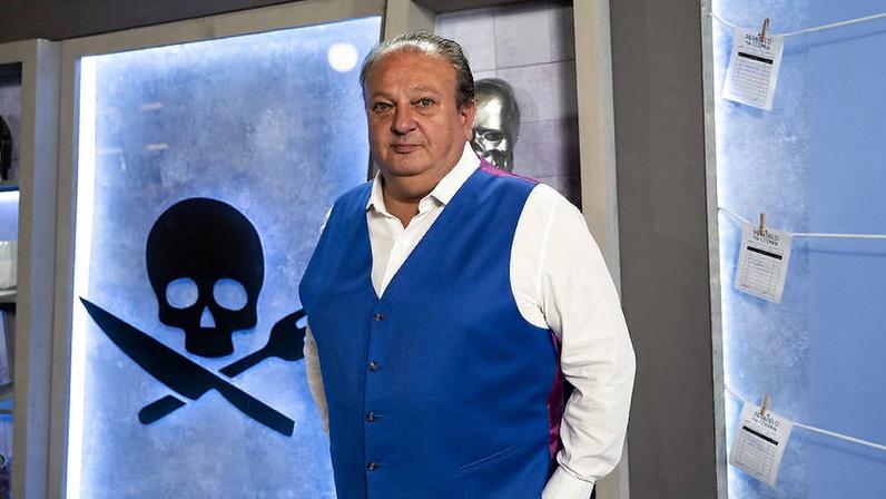 Homem de meia-idade com colete azul e camisa branca está em pé em estúdio. Ao fundo, parede iluminada exibe símbolo de caveira preta com faca e garfo cruzados. Páginas de papel estão penduradas em cordas ao lado direito.
