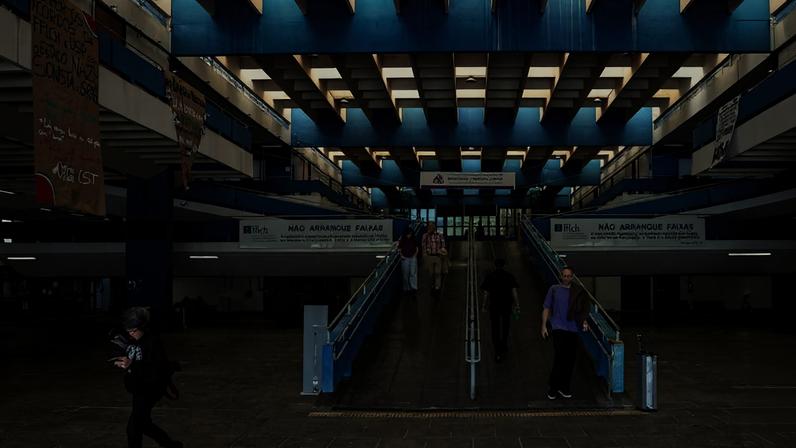 Interior do prédio da reitoria da USP com estrutura azul e teto em padrão quadriculado. Pessoas caminham pela rampa central e pelo piso térreo. Faixas brancas penduradas na parte superior dizem 'NÃO ARREANQUE FAIXAS'. Cartazes de papelão com mensagens manuscritas estão pendurados nas laterais.